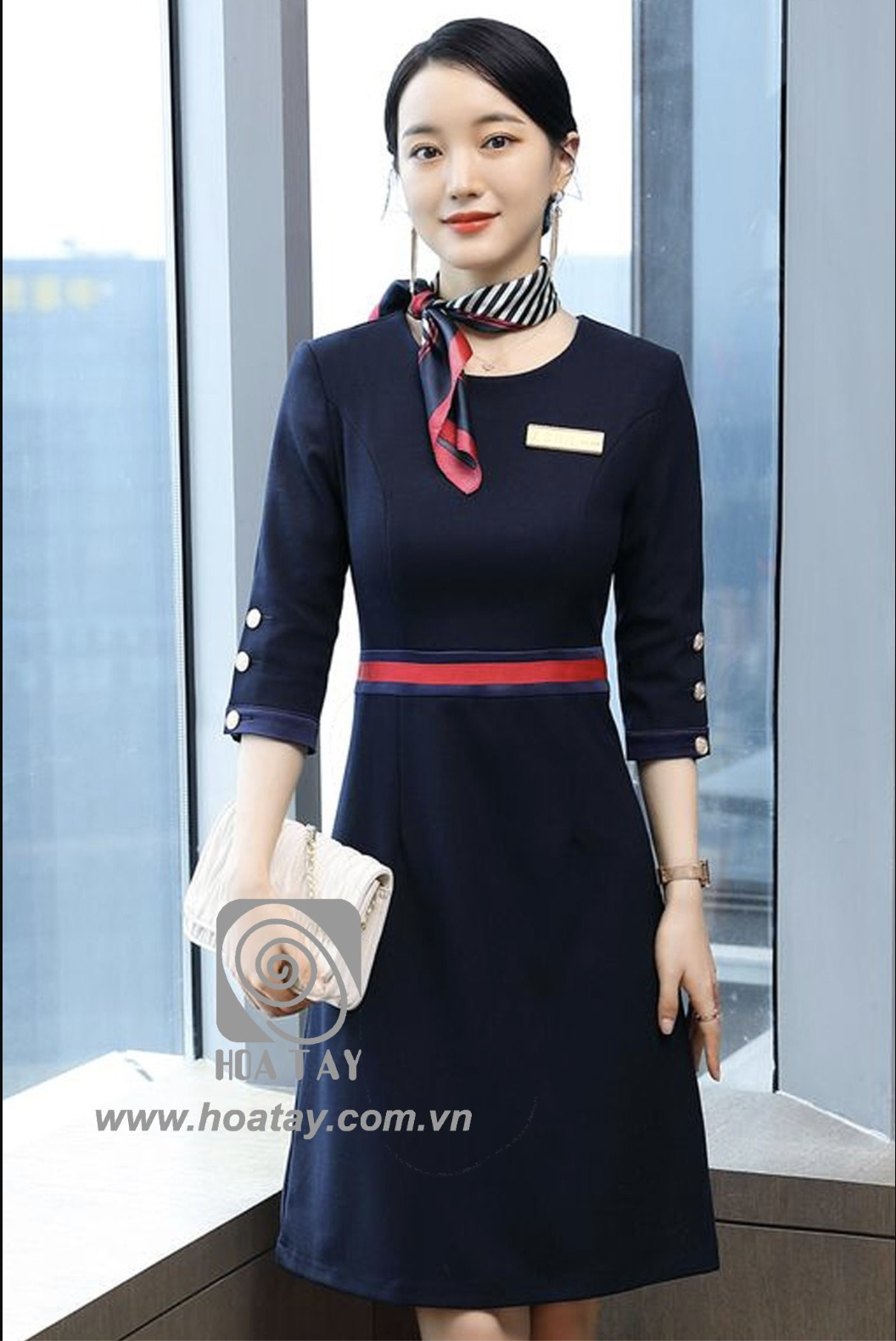 Một nữ nhân viên đứng thẳng, mặc váy liền (đầm) xanh navy, tay lỡ có hàng khuy (nút) trang trí, thắt khăn quàng cổ kẻ sọc, đai eo đỏ, bảng tên kim loại, đứng cạnh cửa sổ kính, ánh sáng tự nhiên.