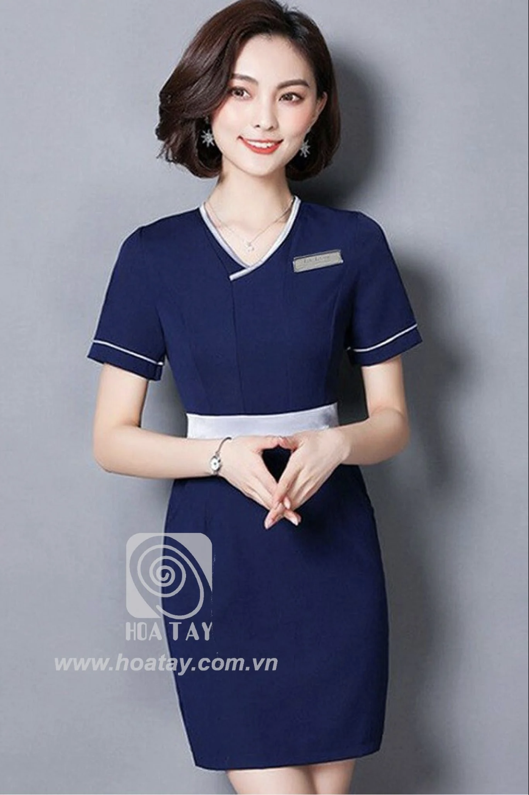 Một nữ nhân viên đang đứng; mặc đồng phục váy liền (đầm) màu xanh navy, cổ chữ V phối viền trắng, tay ngắn, đai eo màu bạc, chất liệu vải đứng form; phông nền xám, ánh sáng studio.