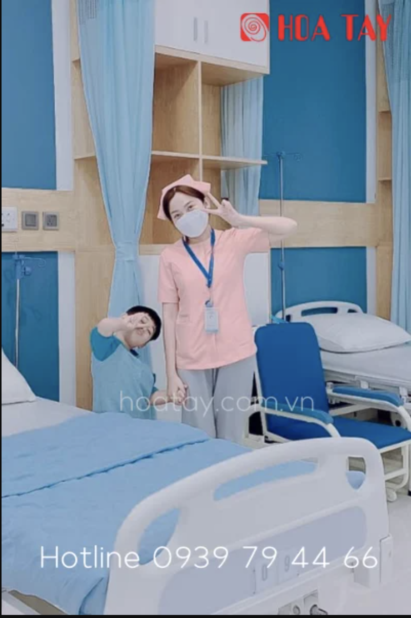 Một nữ điều dưỡng và một bệnh nhi (hai người) đang tạo dáng chữ V; mặc áo blouse hồng cổ tròn (cổ thuyền), quần xám, đội mũ y tá; bối cảnh phòng bệnh nội trú với giường y tế, rèm xanh, ánh sáng trắng 