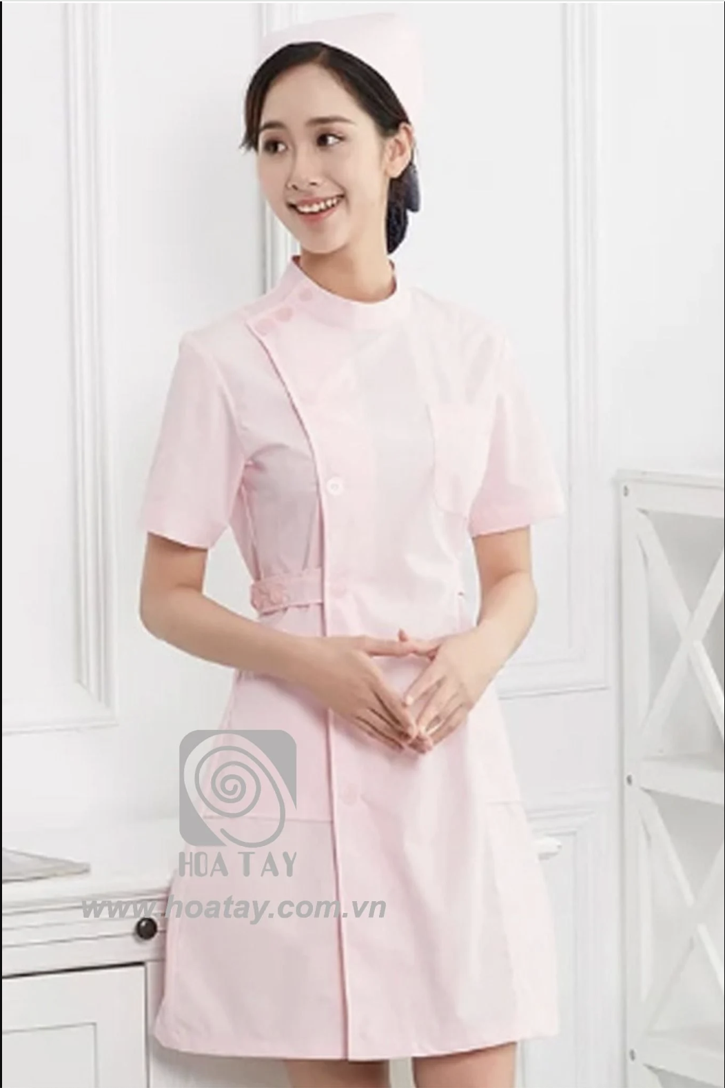 Một nữ điều dưỡng (y tá) mặc đồng phục màu hồng phấn, cổ đứng (cổ tàu), cài khuy lệch (cúc chéo), tay ngắn, chất liệu vải phẳng mịn, kèm mũ y tá cùng tông màu, đứng trong không gian studio sáng trắng.
