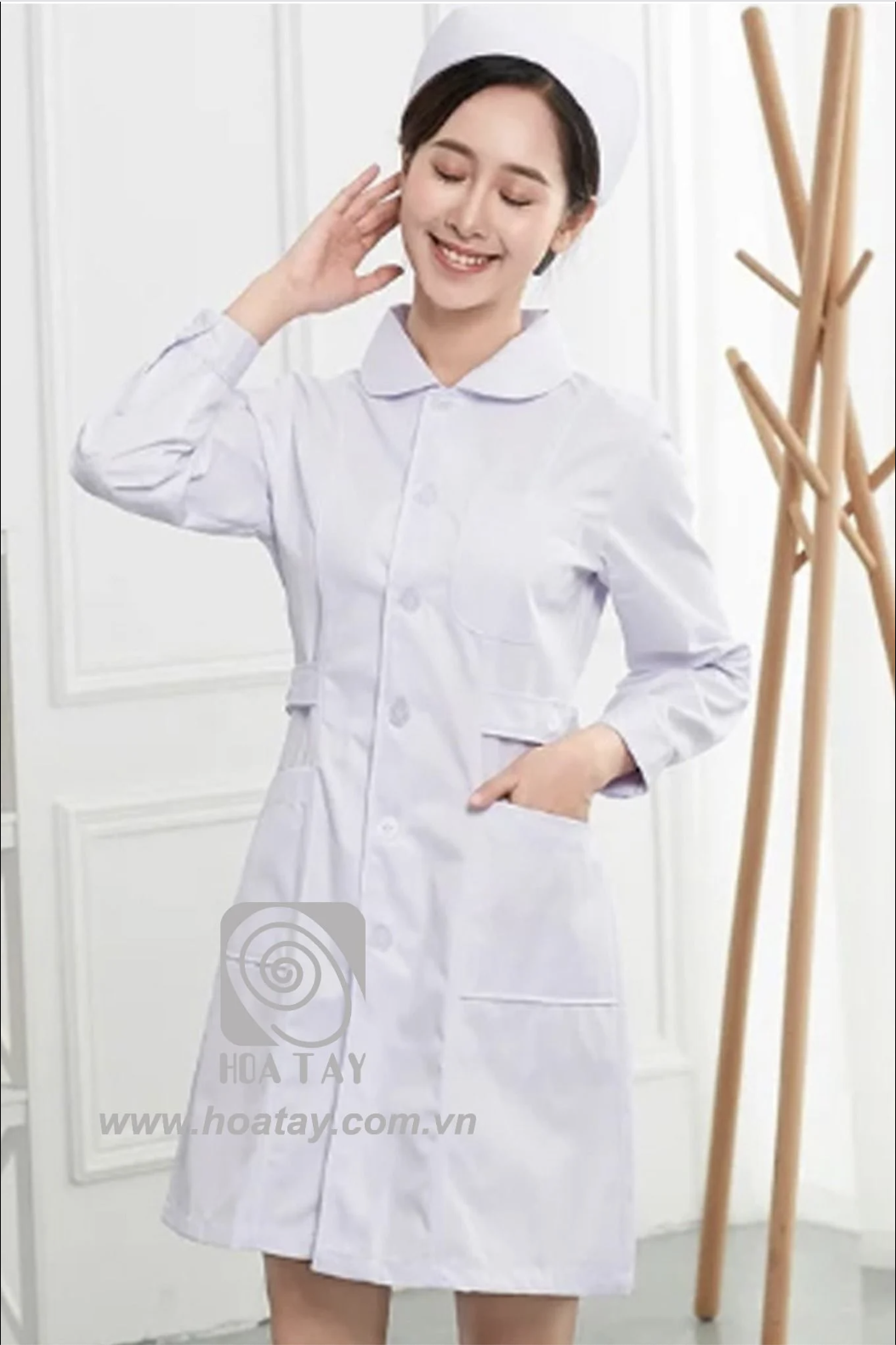 Một nữ y tá đang đứng mỉm cười, mặc áo blouse trắng (áo khoác y tế) dài tay, cổ tròn (cổ Peter Pan), vải cotton, cài cúc dọc thân, có hai túi hông lớn, bối cảnh studio sáng sủa.