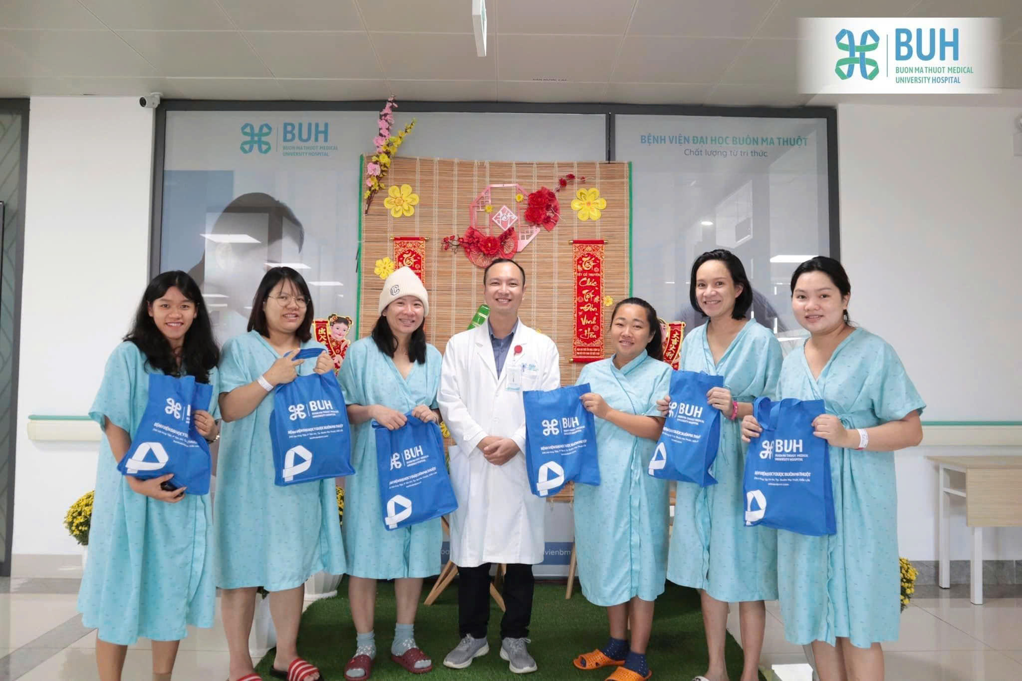 Một bác sĩ nam và sáu bệnh nhân nữ đứng cùng nhau; họ đang cầm túi quà màu xanh dương; bệnh nhân mặc áo choàng bệnh viện (đồ mặc nội trú) màu xanh nhạt, bác sĩ mặc áo blouse trắng; bối cảnh sảnh bệnh