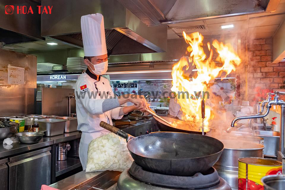 Một đầu bếp nam đang xào nấu; mặc áo khoác trắng cổ tàu (cổ đứng), tay áo dài, chất liệu vải dày dặn, đội mũ toque (mũ cao) trắng; bối cảnh bếp công nghiệp với ngọn lửa bùng lên từ chảo, ánh sáng vàng ấm.