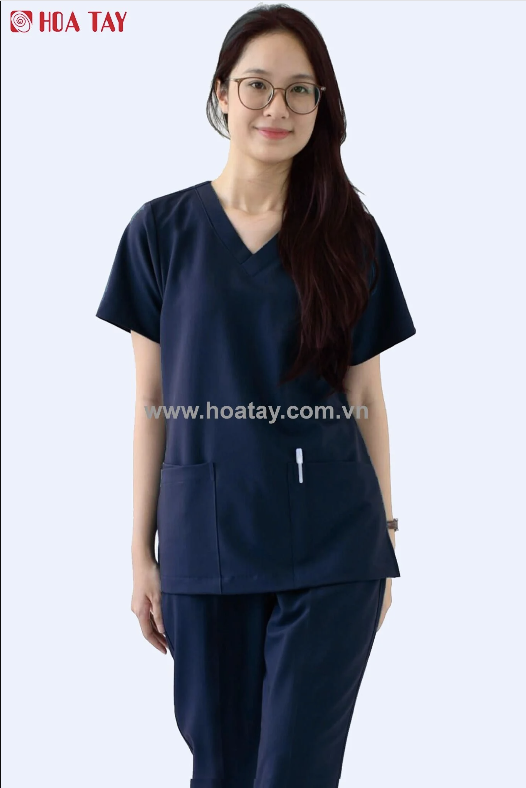 Một nữ nhân viên y tế đang đứng, mặc bộ đồ scrub (đồng phục y tế) màu xanh navy, cổ chữ V, tay ngắn, có hai túi vuông phía trước, chất liệu vải trơn, phông nền trắng, ánh sáng studio.