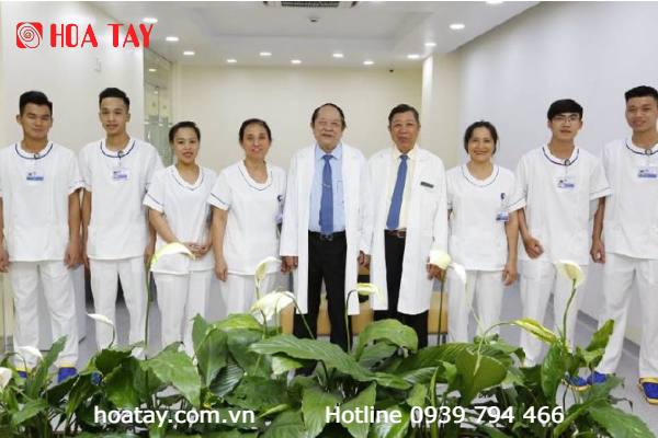 Nhóm 9 người đứng hàng ngang trong phòng khám: 7 nhân viên mặc đồng phục trắng cổ tròn, 2 bác sĩ mặc áo blouse trắng dài. Phía trước là hàng cây hoa trắng. Ánh sáng trần trắng, logo HÒA TÂY ở góc trên